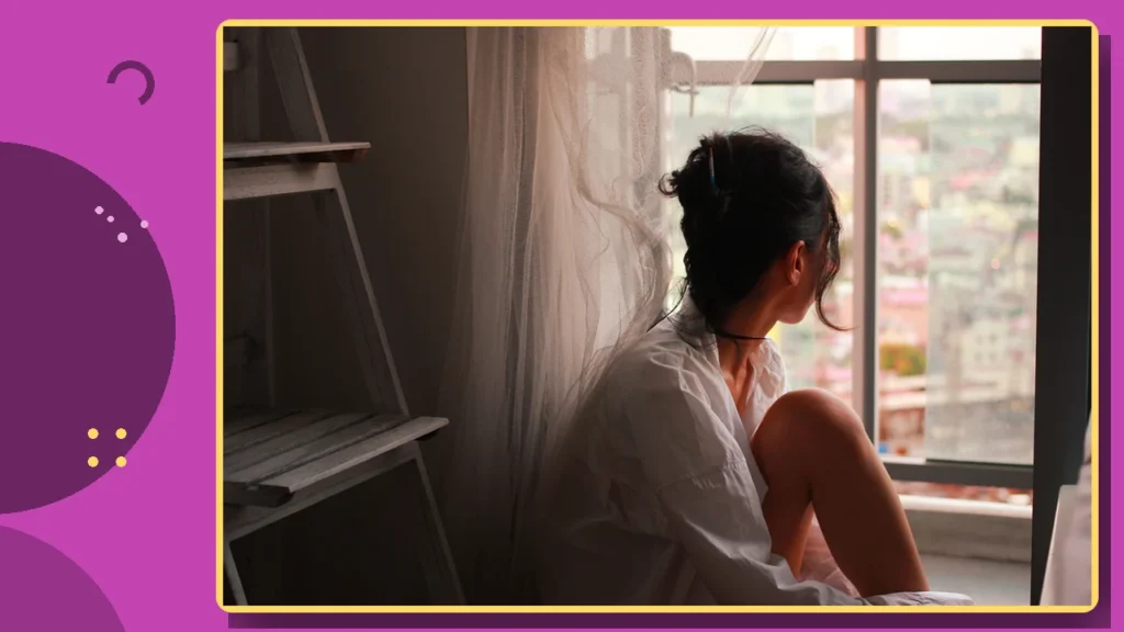 A woman sitting by a window in morning light, looking out thoughtfully.
